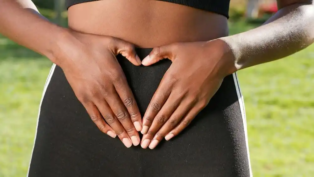 Colonic Irrigation Close-up image of a person forming a heart shape with their hands on their lower back, symbolising colon health and potentially indicating colonic irrigation therapy. The individual is wearing sporty attire with a green backdrop, suggesting a focus on wellbeing and fitness.